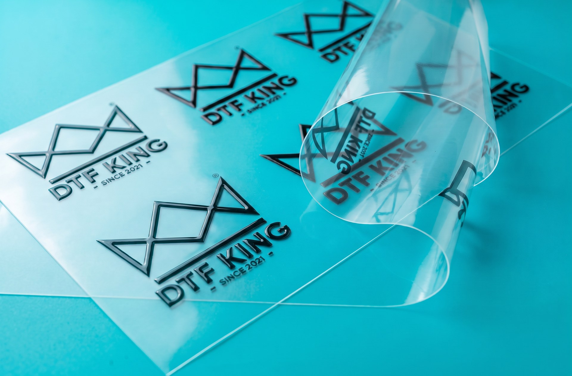 Crystal-clear transparent UV DTF transfer film with translucent printed DTF-King logos on a turquoise background