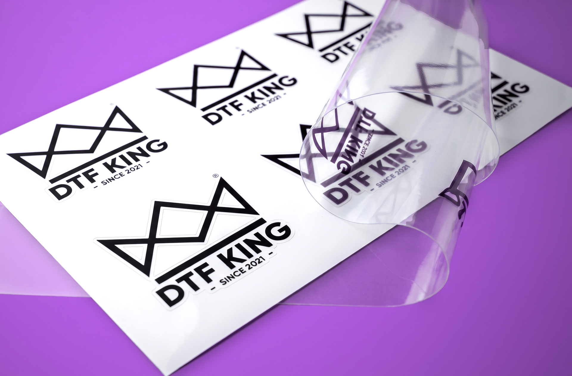 Weiße Vinylfolie mit schwarzen konturgeschnittenen DTF-KING-Logos, darüber eine eingerollte Bahn transparenter Folie