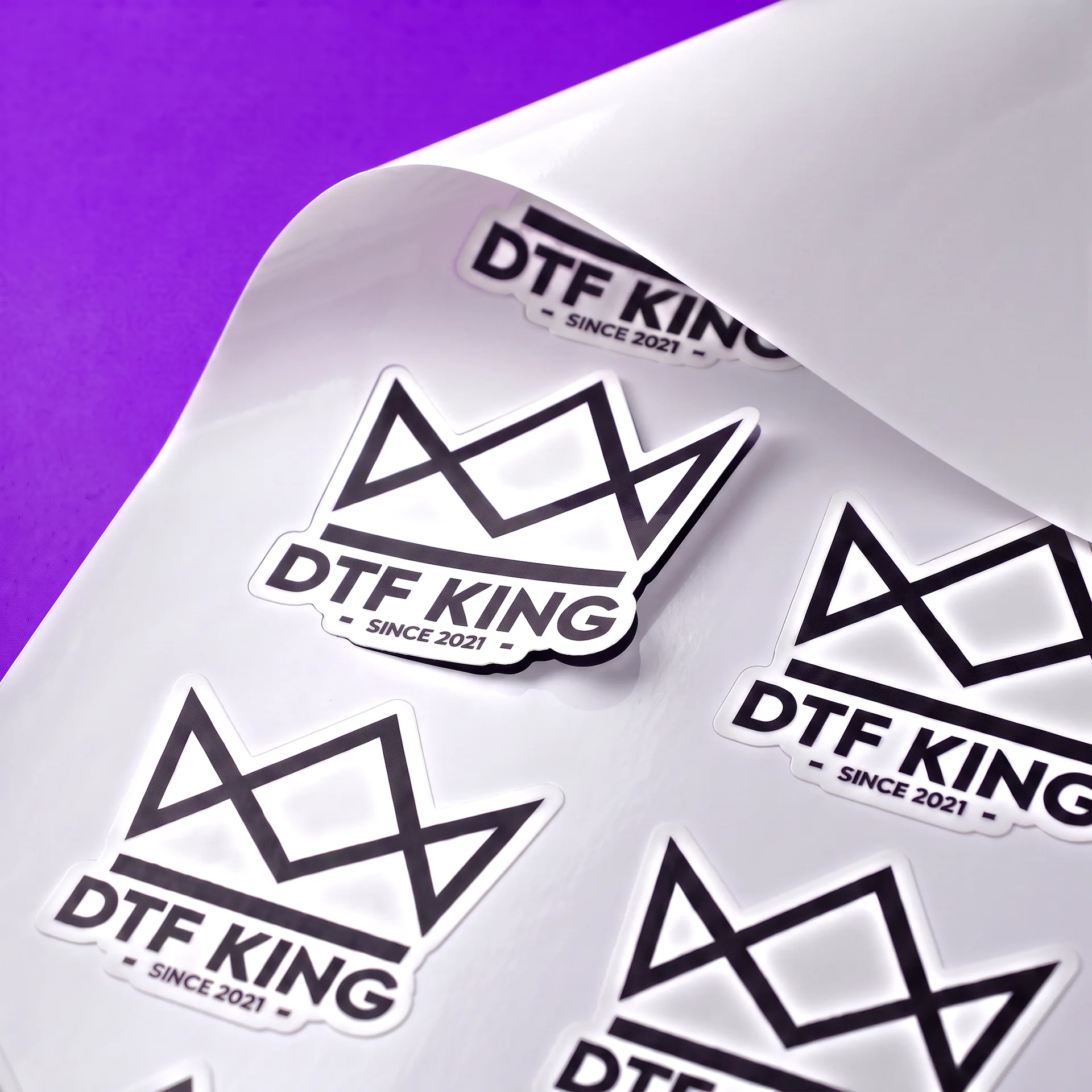 Konturgeschnittene DTF-KING-Vinyl-Sticker auf weißer Trägerfolie, glänzend laminiert, die Folie ist an einer Ecke hochgeklappt