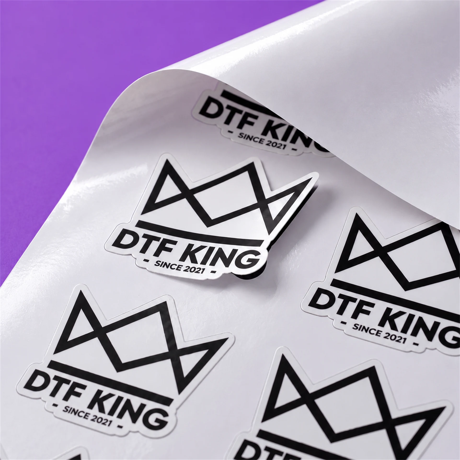 Konturgeschnittene Vinyl-Sticker mit DTF-KING-Logo auf weißer Trägerfolie, glänzend laminiert, ein Sticker liegt lose auf der Bahn